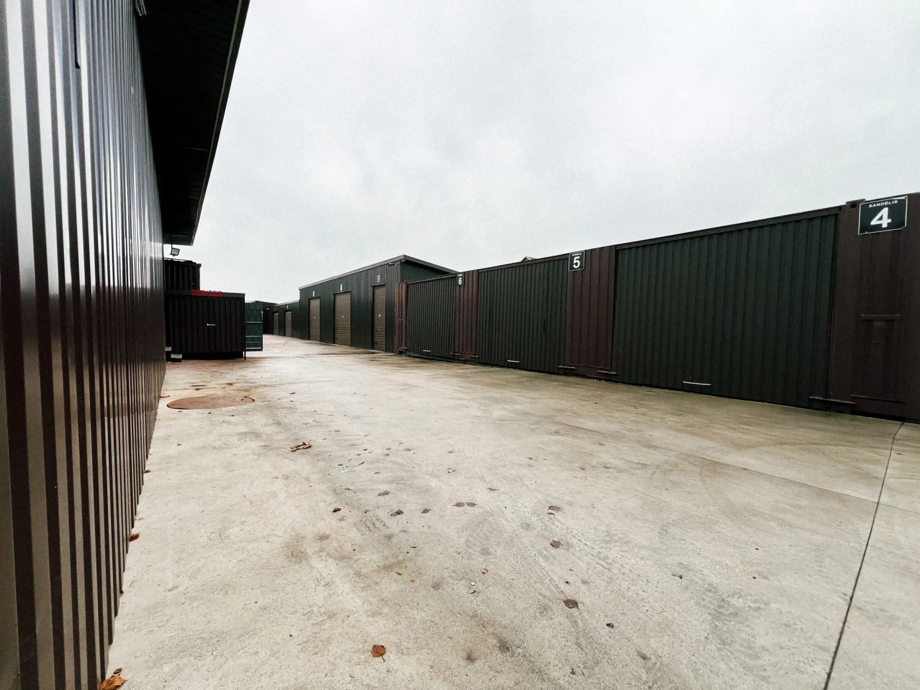 Row of numbered dark green steel storage units with roller doors along a concrete yard at a Gym Factory warehouse site